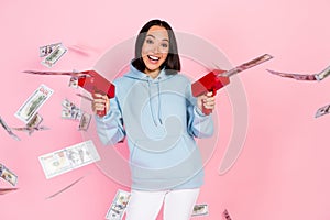 Photo of cute excited lady wear blue hoodie shooting money gun smiling isolated pink color background