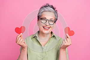 Photo of cute grandmother wear stylish khaki shirt holding two paper red postcards heart valentine day isolated on pink