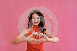 Photo of a cute optimistic curly brown middle-aged woman in a red dress against a pastel pink background. She smiles and