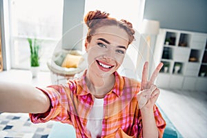 Photo of cute positive girl toothy smile show v-sign take selfie enjoy free time modern apartment indoors