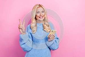 Photo of cute positive woman blue sweater hold phone show v-sign instagram telegram whatsapp facebook isolated on pink