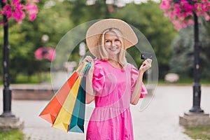 Photo of cute pretty lady pensioner dressed pink clothes headwear walking holding shopping bags credit card smiling