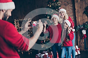 Photo of daddy making photo of brother sister and mommy showing v-sign near decorated newyear tree indoors x-mas spirits