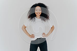 Photo of delighted curly woman keeps both hands on waist, smiles gently, has slim figure, wears white t shirt and black jeans,