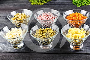 Ingredients shredded for salad in transparent plates on a black wooden background. Olives, pickled cucumbers, sausages, carrots, p