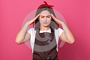 Photo of depressed exhausted young woman with black hair being under stress, having headache, putting hands on temples, closing