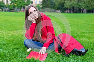 Photo of depressed passive sad teen girl sitting on green grass lawn is not willing to do home task looking away leaning on hand