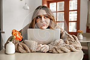 Photo of disappointed woman in sweater looking aside with suspicious gaze, while hugging laptop in coffee shop