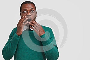 Photo of dismayed frightened black hipster covers mouth with both hands, wears green outfit, keeps eyes popped out photo