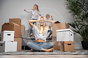 Photo of displeased woman sitting on floor among cardboard boxes and boy, girl jumping on sofa