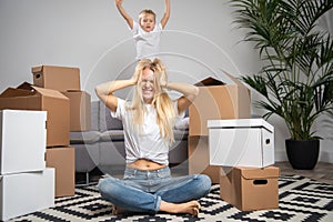 Photo of displeased woman sitting on floor among cardboard boxes and boy jumping on sofa