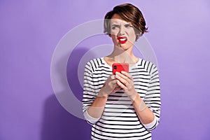 Photo of distrustful girl hold telephone grimacing look empty space wear striped shirt isolated purple color background