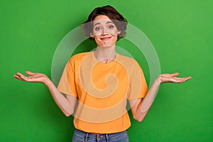 Photo of doubtful funny young woman dressed orange t-shirt shrugging shoulders isolated green color background