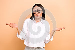 Photo of doubtful unsure woman dressed white shirt eyewear shrugging shoulders isolated beige color background