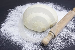A photo of dough, rolling pin and wheat white flour sprinkled on black wooden table. Selective soft focus. Dough ready for cooking