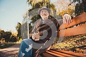 Photo of dreamy inspired grey hair old man enjoy rest relax october autumn park sit bench drink takeaway coffee mug wear