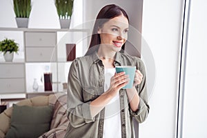 Photo of dreamy pretty young woman dressed green shirt looking window drinking coffee indoors house home room