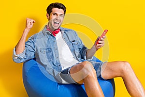 Photo of ecstatic funky guy wear jeans jacket shorts sit on pouf hold smartphone win betting isolated on bright yellow