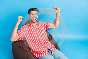 Photo of ecstatic man with bristle dressed striped shirt sit on bean bag scream yes win lottery fists up isolated on