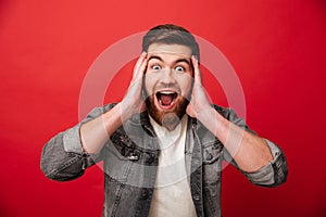 Photo of emotional guy 30s wearing beard in jeans jacket screaming and grabbing head in delight, isolated over red background