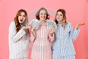 Photo of envious women 20s in colorful striped pajamas expressing irritation while lucky female friend holding lots of money doll