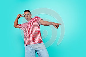 Photo of excited cute young guy dressed red t-shirt dancing pointing empty space showing v-sign isolated turquoise color