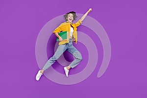 Photo of excited funny young woman dressed yellow shirt jumping high holding copybooks rising fist  purple color