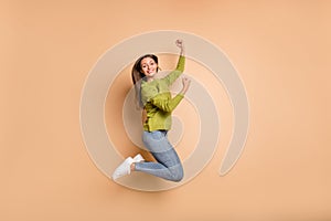 Photo of excited lady jump raise fists wear green pullover jeans footwear isolated beige color background