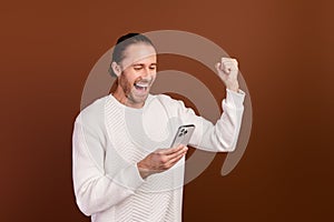 Photo of excited lucky guy dressed white pullover rising fist winning game modern device isolated brown color background