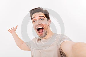 Photo of excited man in casual t-shirt and bristle on face screaming in happiness while taking selfie, isolated over white