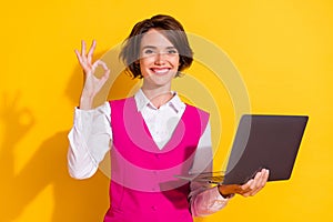 Photo of excited shiny young lady wear pink suit holding modern device showing okey smiling isolated yellow color