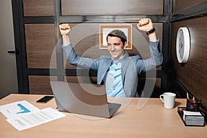 Photo of excited young manager working with laptop and business documents celebrating work success in modern office.