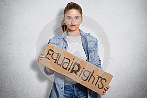 Photo of feminist in denim jacket, holds sign written Equal rights, shows that women have rights and power, isoated over white con