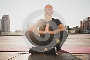 Photo of focused mature old man sit fitness mat hold hands together close eyes meditate wear t-shirt urban town outdoors