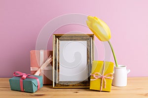Photo frame with gift boxes and yellow tulip on wooden table with pink background