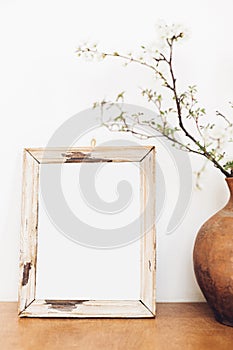 Photo frame mockup and blooming cherry branches in vase on wooden table against white wall. Empty picture frame template and