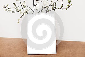 Photo frame mockup and blooming cherry branches on wooden table against white wall. Empty picture frame template and spring