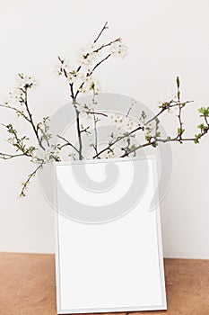 Photo frame mockup and blooming cherry branches on wooden table against white wall. Empty picture frame template and spring