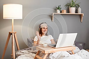 Photo of friendly delighted attractive woman wearing sleeping mask having video call on laptop while lying on bed, waving hand,