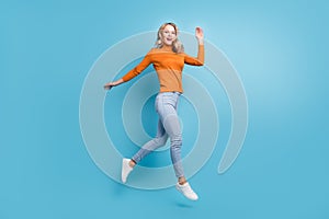 Photo of friendly pretty young lady wear orange sweater jumping high waving arm smiling isolated blue color background