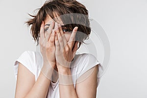 Photo of frightened woman with short brown hair in basic t-shirt keeping hands over her face and looking at camera