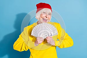 Photo of funky shiny mature lady wear yellow hoodie red headwear holding money fan smiling isolated blue color