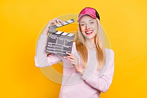 Photo of funny cute young lady wear pink sweater headwear smiling holding clapper board isolated yellow color background