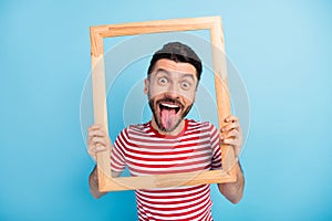 Photo of funny fooling young guy wear red outfit holding looking wooden frame showing tongue isolated blue color