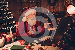 Photo of funny santa claus look pc screen direct finger laugh joke wear hat pullover in north pole office indoors