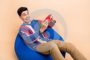 Photo of funny young handsome brunet hair male sit bean bag playing mobile games download new update isolated on beige