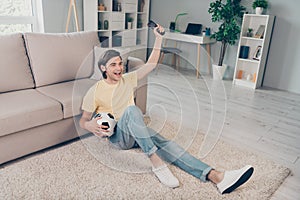 Photo of glad excited astonished football fan guy sit carpet hold ball celebrate victory wear yellow t-shirt home