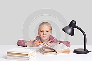 Photo of good looking small child hold pencil, looks at camera with blue eyes, reads different books for having good knowledge, po