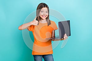 Photo of good mood adorable little girl wear orange stylish t-shirt holding laptop show thumb up isolated on teal color