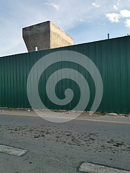 Photo of green zinc fence on asphalt road and partially concrete pillar in the morning